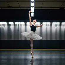 The image captures a dynamic scene inside a subway station where a ballerina is frozen in a mid-motion pose. The ballerina, dressed in a black leotard and ballet shoes, stands out against the urban backdrop. She's caught mid-pointe with her left leg exten